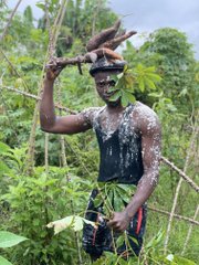 A man standing in a lush green forest, covered in white substance, wearing a leafy headpiece and skirt while holding a stick above his head.