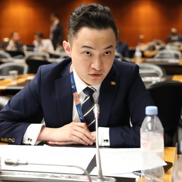 A man in a dark blue suit and striped tie sits at a conference table with a microphone and water bottle.