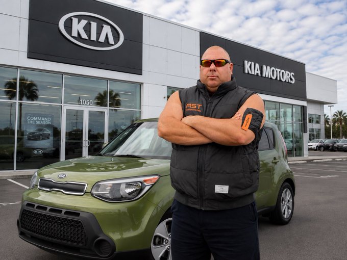 A man wearing an "AST" branded vest and sunglasses stands with crossed arms in front of a Kia Motors dealership, next to a green Kia Soul, with the post celebrating his first Kia Soul purchase as a memorable achievement.