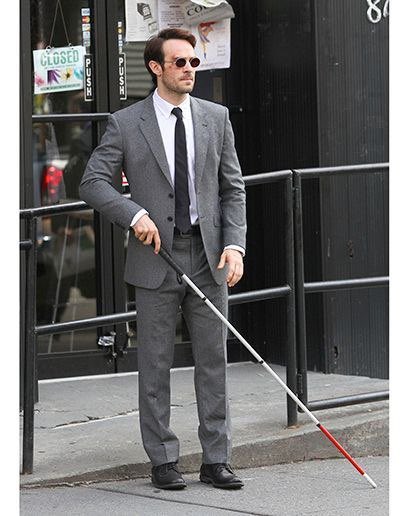 A man in a gray suit, white shirt, black tie, and sunglasses stands on a sidewalk holding a white cane with a red tip, indicating he is blind.