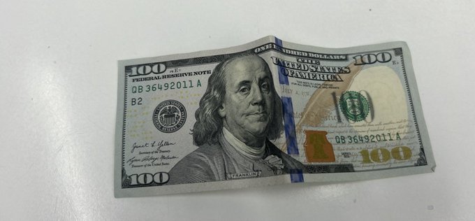 A slightly worn US $100 bill featuring Benjamin Franklin lying flat on a light-colored surface, with visible serial numbers DB 36492011 A.