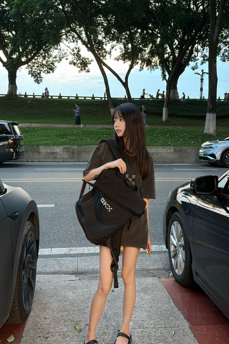 A young woman in an oversized dark "OKX" t-shirt dress stands between two parked cars on a tree-lined street, holding a matching OKX checkered-pattern shoulder bag as she poses for a casual weekend photo. The branded merchandise reflects the post's promotion of OKX merch during a leisurely outing, set against a scenic backdrop of greenery and passersby.