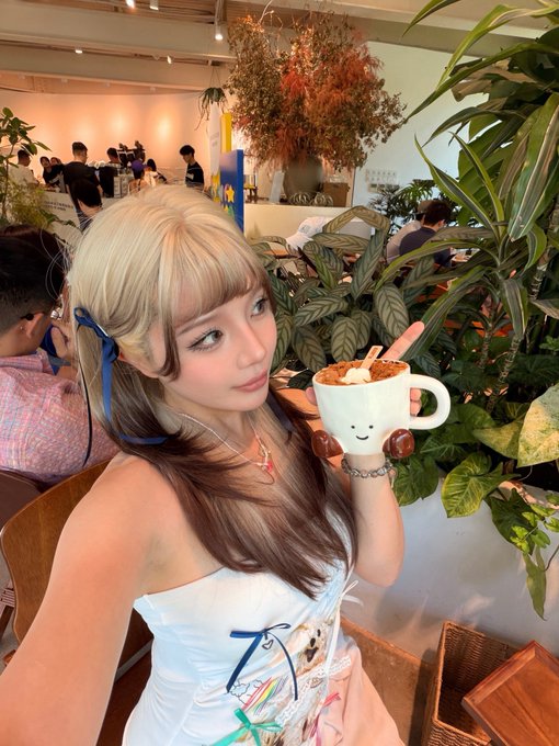 The image features a woman with blonde hair wearing a white dress, holding a cup of coffee that has a smiley face drawn on it. The background includes several potted plants, creating an atmosphere of relaxation or leisure. There are also other people in the scene, but they do not seem to be the main focus of the photo.