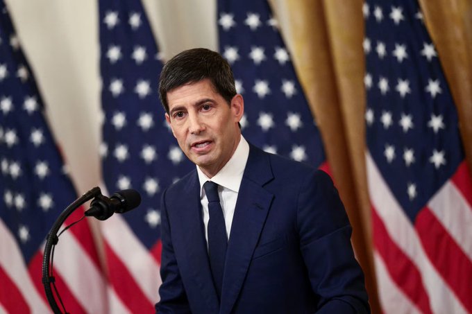 A man in a dark blue suit and tie speaks at a microphone in front of multiple American flags.