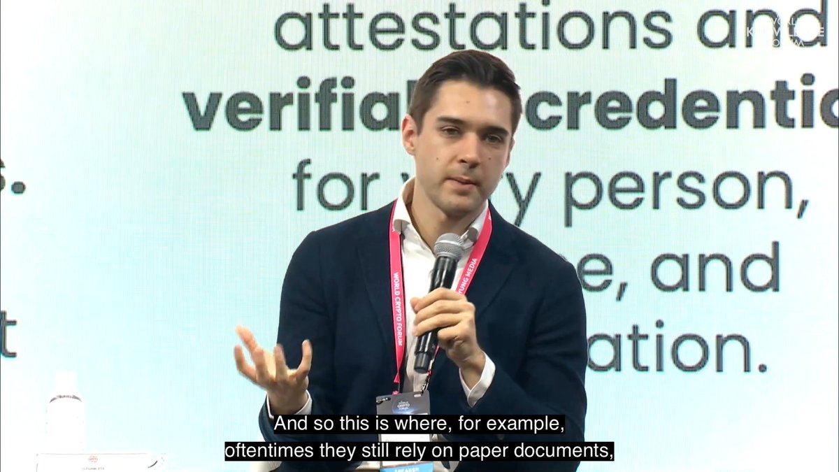 A young man in a dark blazer speaks into a microphone at the World Economic Forum, gesturing with his left hand. The screen behind him displays partial text about "attestations" and "verifiable credentials," while subtitles read, "And so this is where, for example, oftentimes they still rely on paper documents." The image illustrates Dom Schiener's discussion about a UK pilot solution that reduced customs clearance time from days to hours.