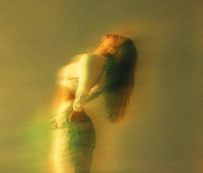 A woman with long hair arches her head back in a hazy, golden-yellow lit scene, embodying the introspective and emotional mood of the post's message about the complexities of life. She wears a cropped white long-sleeve top and light jeans, with soft light flares adding to the dreamlike atmosphere that reflects the post's themes of self-reflection and existence.