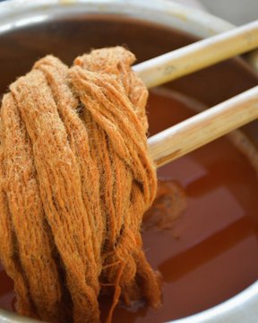 A close-up photo of chopsticks lifting fibrous, brown-colored food (resembling textured meat or tripe) from a dark, reddish-brown broth.