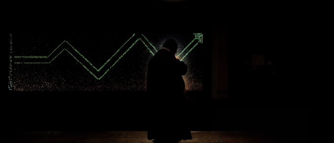 A dark, atmospheric image showing the silhouette of a person sitting in front of a window displaying a bright green line graph with an upward arrow trending on a black background.