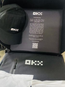 A photograph of OKX merchandise including a black OKX branded cap alongside black packaging boxes featuring OKX branding and a QR code.