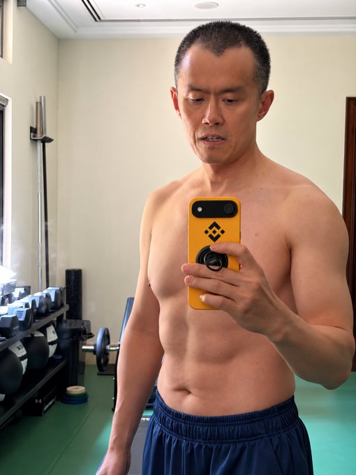 A shirtless man takes a mirror selfie with a yellow smartphone in a gym setting, with dumbbells and equipment visible in the background. The image visually ties to the post's concept of a "#BNB Phone," showing a person holding a branded-looking phone as if previewing the hypothetical product.