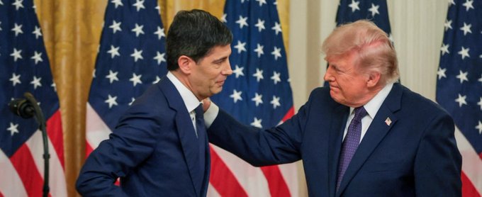 Two men in dark suits stand in front of American flags; one has his hand on the shoulder of the other in what appears to be a formal political setting.