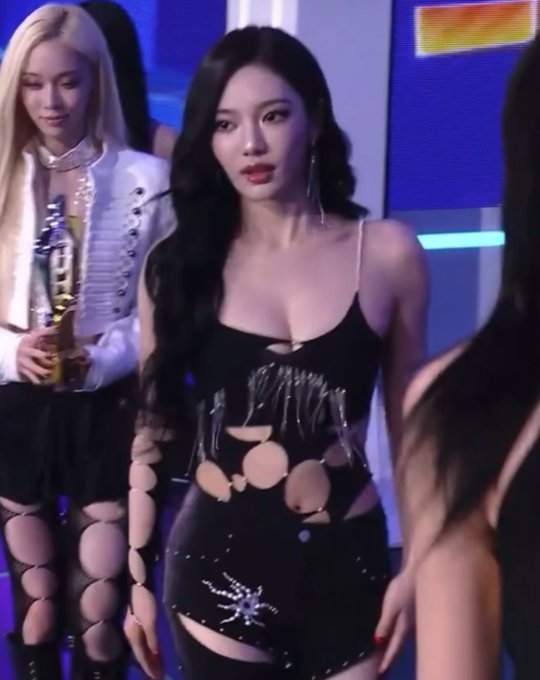 A performer with long dark wavy hair stands on stage in a black cutout bodysuit embellished with rhinestones, with other performers visible in the background.