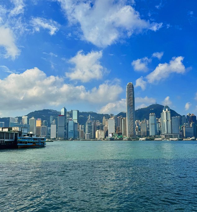 A daytime view of a city skyline across a body of water, featuring tall skyscrapers and mountains in the background under a blue sky with scattered clouds.