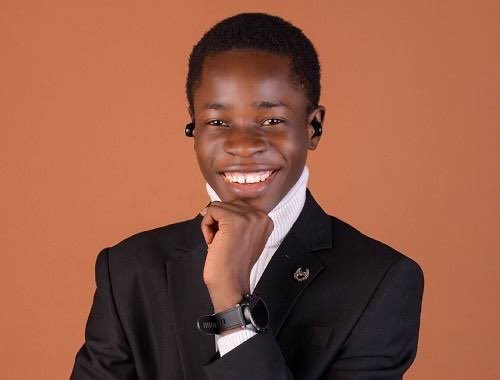 A smiling young man wearing a black suit, white shirt, and black earbuds poses against a brown background with his hand near his chin.