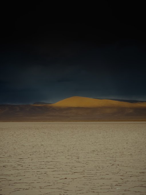 A moody landscape photograph showing a flat, cracked desert floor stretching toward a distant mountain under a dark, dramatic stormy sky.