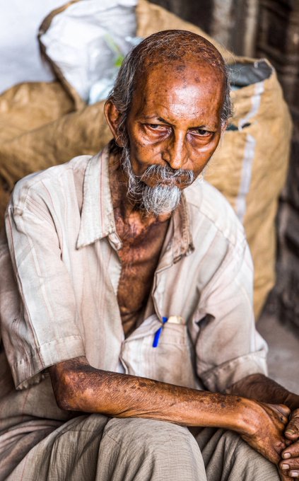 Portrait of an elderly bearded man with weathered, sun-darkened skin wearing a worn beige shirt with a blue pen in the pocket, sitting with what appears to be sacks behind him.