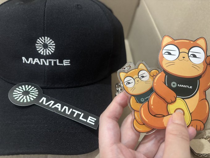 A hand holds up two orange cartoon cat keychains branded with 'MANTLE' logo, displayed next to a black MANTLE baseball cap.