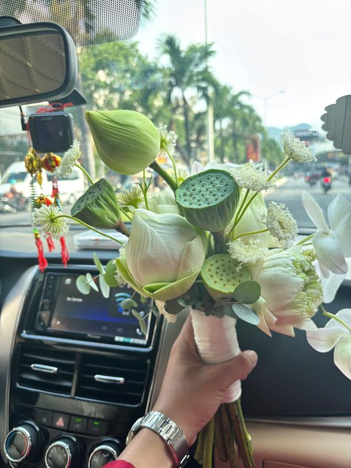 A hand wearing a watch holds a bouquet of white lotus flowers and lotus pods inside a car with a flower-shaped air freshener hanging from the rearview mirror.