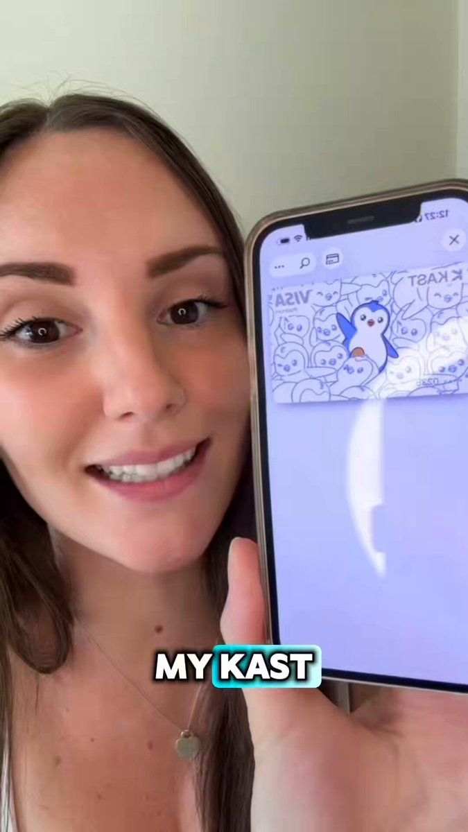 The image shows a woman holding up her phone displaying an image of a penguin card with a message that reads "My KAST". The penguin card has a picture of a fire, which might be a part of a game or a challenge related to the card. The woman is smiling and appears to be excited about the penguin card on her phone screen.