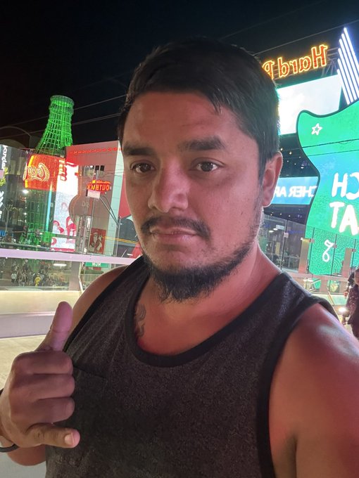 The image features a man with a beard, wearing a tank top, standing outside at night. He is giving a thumbs up gesture while posing for the photo. The background of the image shows a city street scene with neon signs and buildings lit up at night. There are also some bottles visible in the scene. The post that accompanies this image reads: "OG @PumpfunEco test coin". This suggests that the man is promoting or showcasing an online store called Pumpfun Eco, which sells coins related to urban environments and city life.
