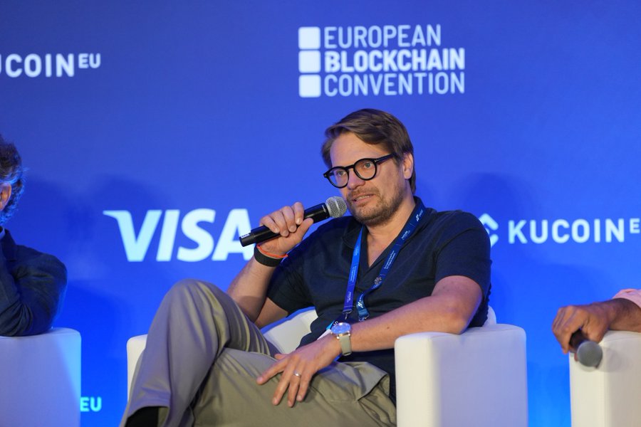 A man with glasses and a beard sits in a white chair holding a microphone at the European Blockchain Convention, with 'VISA' and 'KUCOIN.EU' logos visible on the blue backdrop.