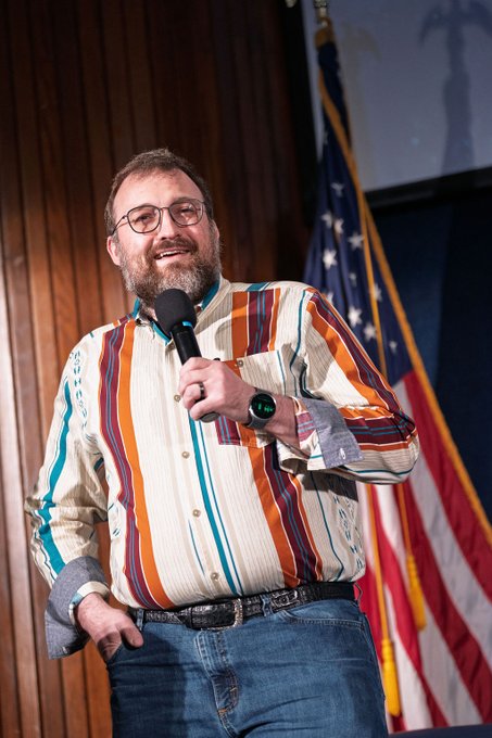 A man with glasses, a beard, and a colorful striped shirt holds a microphone while performing or speaking on a stage with an American flag behind him.