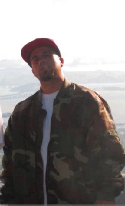 A portrait of a man with a goatee wearing a red baseball cap backwards and a camouflage jacket over a white t-shirt, with a body of water and land visible in the background.