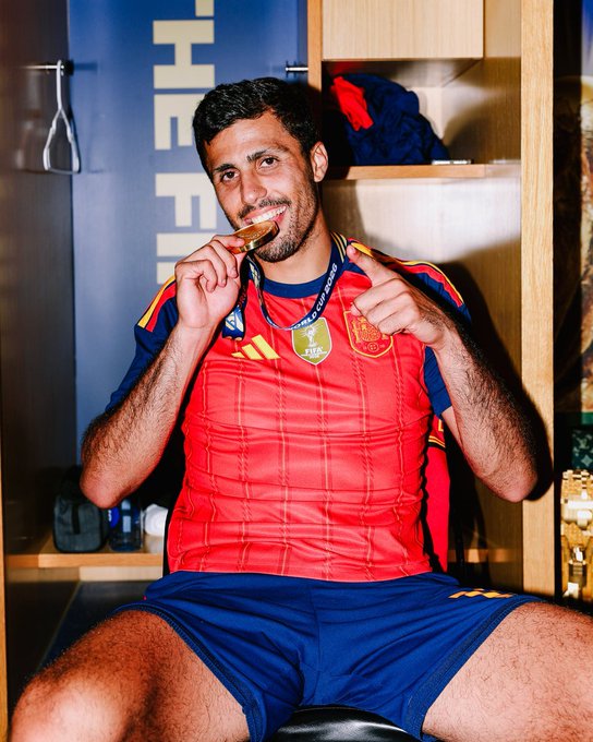 A smiling soccer player in a red Spain national team jersey sitting in a locker room and biting a gold medal.
