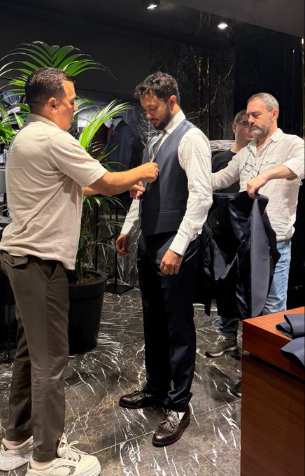 An indoor scene inside a high-end men's tailoring shop, where a tailor in a beige polo shirt assists a customer wearing a white shirt, grey vest, and dark trousers, while another man stands behind them holding a dark jacket.