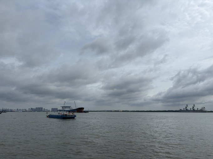 A small blue and white passenger boat navigates calm waters under an overcast sky, with a city skyline and port cranes visible in the distance.