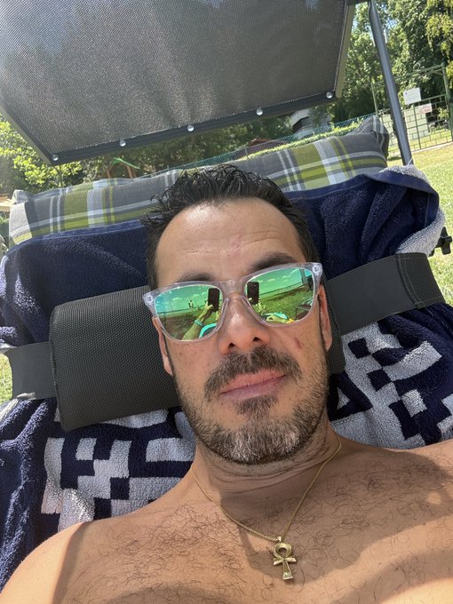 A shirtless man wearing green reflective sunglasses and an ankh necklace lies on a lounge chair outdoors.