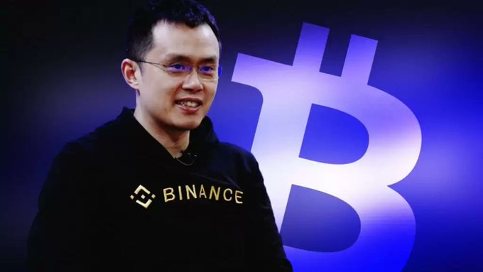 A man wearing glasses and a black Binance hoodie smiles in front of a large blue illuminated Bitcoin logo.
