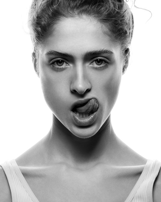 A black-and-white portrait of a young woman with her hair pulled back, looking directly at the camera with her tongue slightly out.