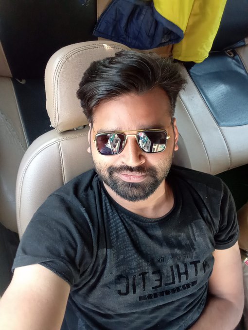 A bearded man wearing sunglasses and a black "ATHLETIC" t-shirt takes a selfie inside a vehicle, fitting the post's "GM CT from India" greeting as a casual, on-the-go check-in while he highlights his interest in the $RIVER ecosystem and Tria platform.
