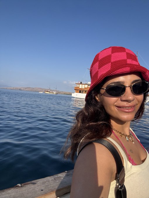 A selfie of a smiling woman wearing a pink checkered bucket hat and oval sunglasses, with boats visible on the water in the background on a sunny day.