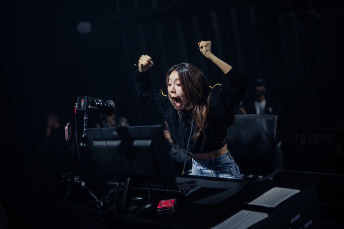 A young woman with dark hair in a black sweater and jeans stands at a computer setup with multiple monitors, throwing her fists in the air and yelling enthusiastically in a dark environment.