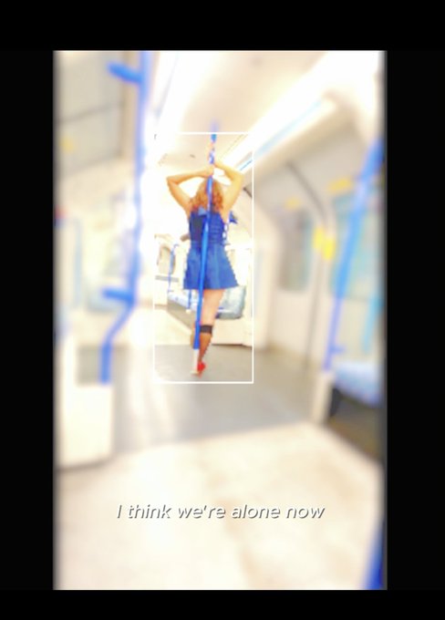 An edited image of a woman in a blue dress standing on a subway or train, captured from behind. The photo is framed within a white border and bears the caption 'I think we're alone now' at the bottom.