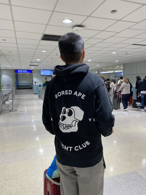 A person stands in what appears to be an airport terminal, wearing a black hoodie with a "Bored Ape Yacht Club" skull logo on the back.
