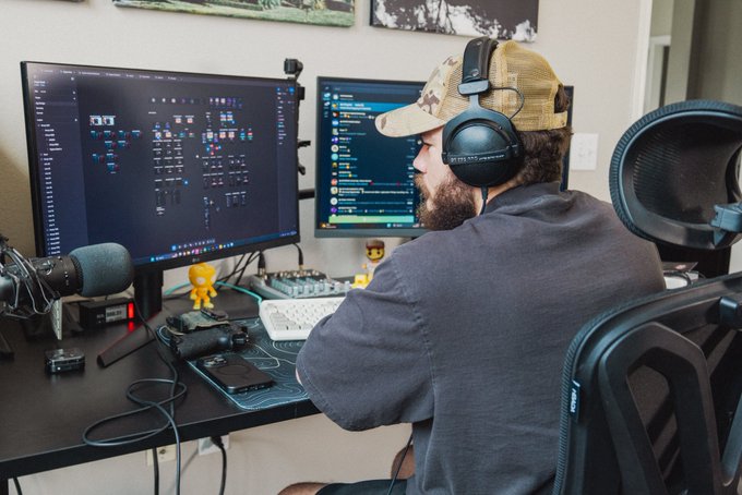 A bearded designer wearing headphones and a cap works at his dual-monitor setup, with one screen displaying design software and the other showing what appears to be a chat interface, highlighting his hands-on creative workflow behind the accomplishments mentioned in his post.