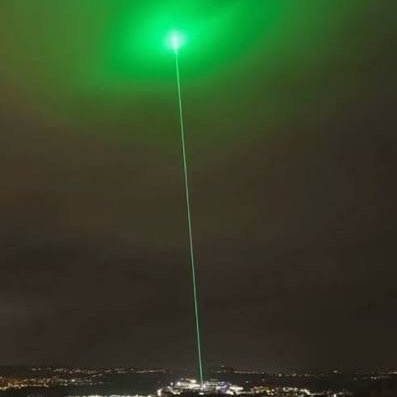 A powerful green laser beam pierces the night sky above a city, illuminating the atmosphere with a bright glow. The image visually amplifies the post's bullish crypto sentiment, symbolizing Bitcoin's unstoppable upward trajectory as it "shoots to the moon," mirroring the surge past market fears and bearish headlines.