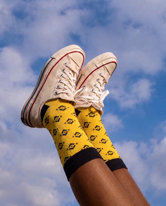Two legs wearing yellow patterned socks and cream-colored Converse high-top sneakers stretch up against a bright blue sky with scattered clouds, embodying the carefree spirit of getting outside and enjoying the day.