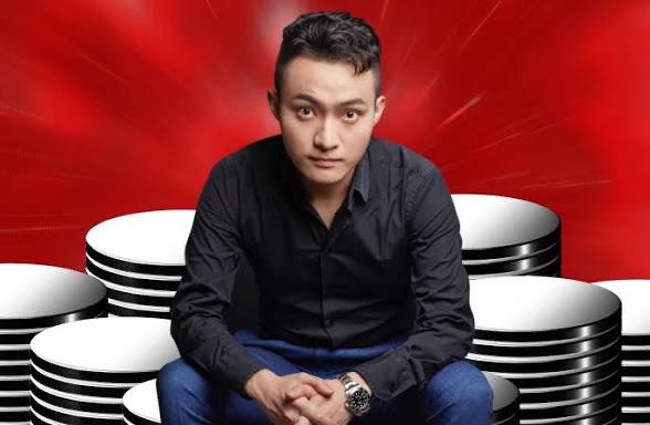 A man in a black button-up shirt and jeans sits with his hands clasped in front of stacked poker chips against a red background.
