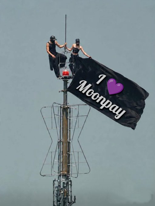 Two people in black outfits on a tower structure flying a black flag with 'I ❤ Moonpay' written in white text.