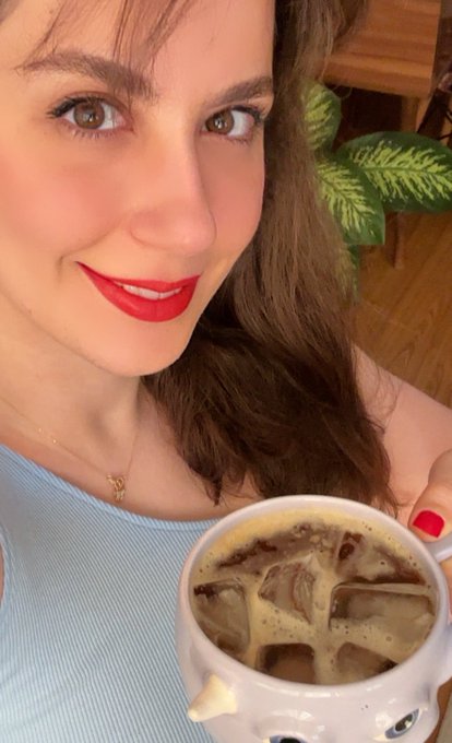 A close-up selfie of a woman with brown hair, wearing red lipstick and a light blue top, holding a white cat-shaped mug filled with iced coffee.