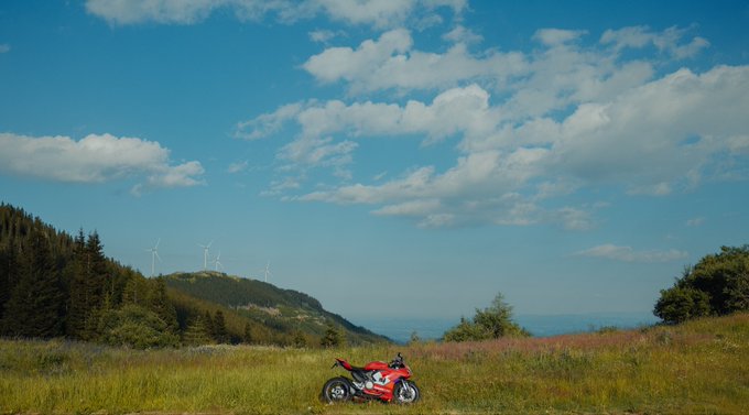 A red Ducati motorcycle is parked in a grassy field with rolling hills, trees, and wind turbines in the background, a perfect setting for reflecting on life and engineering.