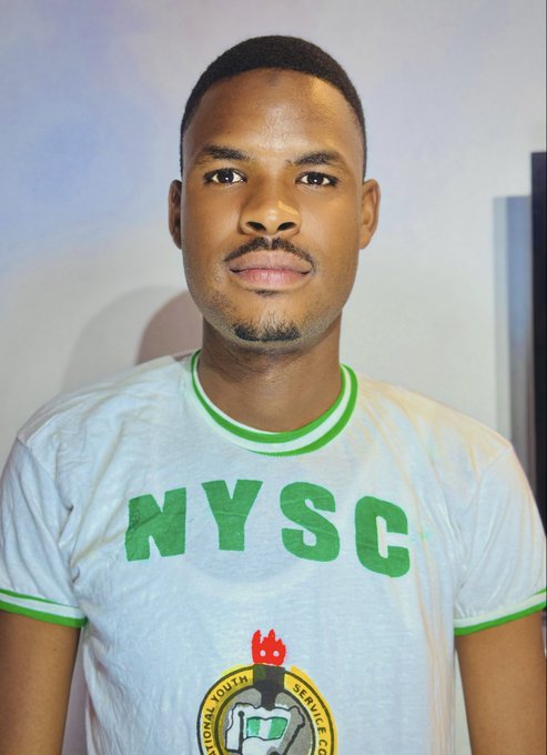 A portrait photo of a young man wearing a white NYSC (National Youth Service Corps) t-shirt with green trim, featuring the official NYSC logo with a torch and handshake emblem.