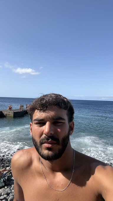 A bearded man takes a shirtless selfie on a rocky beach with blue ocean waves and a stone pier in the background, while the accompanying post asks followers to "Drop your $SOL address" — suggesting he's greeting the crypto community with a morning message.