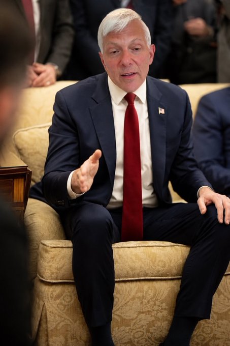 A man with white hair wearing a dark suit and red tie with an American flag pin, seated on a beige couch and gesturing while speaking.