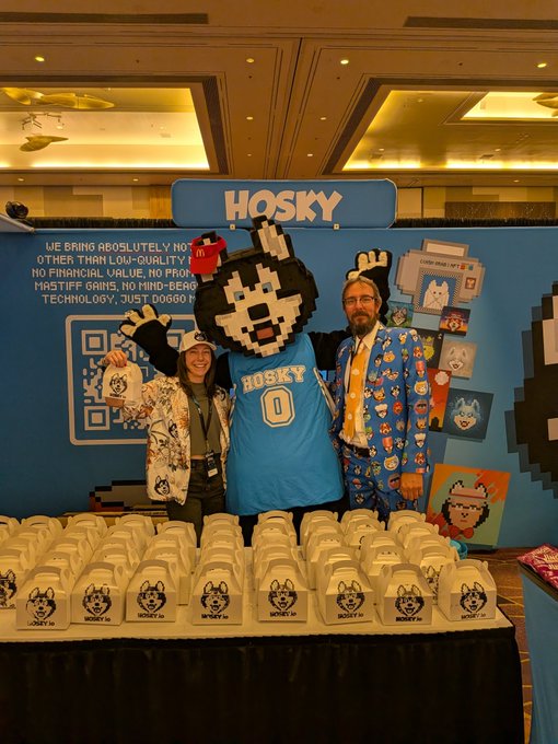 Two people pose with a Hosky mascot in a blue "HOSKY" jersey (number 0) at a convention booth featuring rows of white takeout boxes printed with the Hosky dog logo, alongside a banner reading "WE BRING ABSOLUTELY NO[THING] OTHER THAN LOW-QUALITY... NO FINANCIAL VALUE, NO PROFIT... TECHNOLOGY, JUST DOGGO!"—capturing the community-focused energy mentioned in the caption at the Rare Evo event.