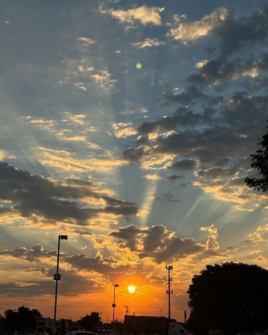 A scenic sunset photo capturing sunbeams (crepuscular rays) radiating through scattered clouds above a parking lot, with silhouettes of light poles, trees, and vehicles against an orange and blue sky.
